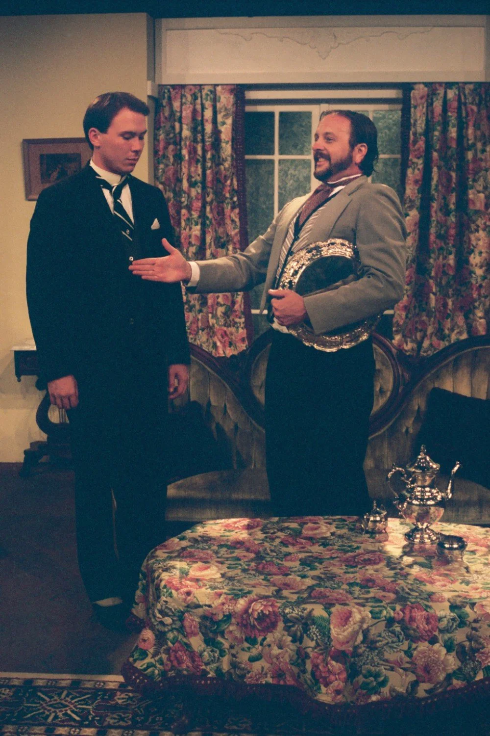 Two actors in a stage play, one wearing a black suit and the other a gray suit, in a vintage-style living room set with a floral tablecloth and decorative items.