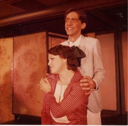 A smiling couple, with a woman sitting in front wearing a red-striped jacket and a white shirt, and a man standing behind her in a light-colored striped suit with a large bow tie, both in a warmly-lit room.