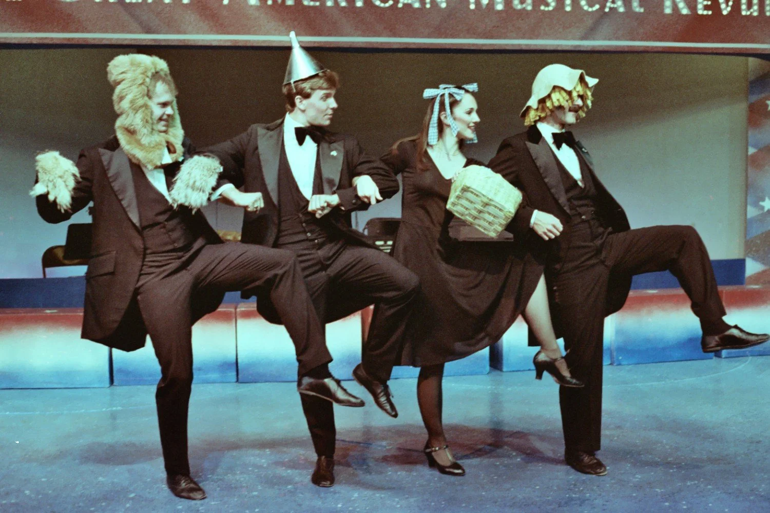 Four people in formal wear, adorned with costumes resembling characters from 'The Wizard of Oz,' perform a dance routine on stage.