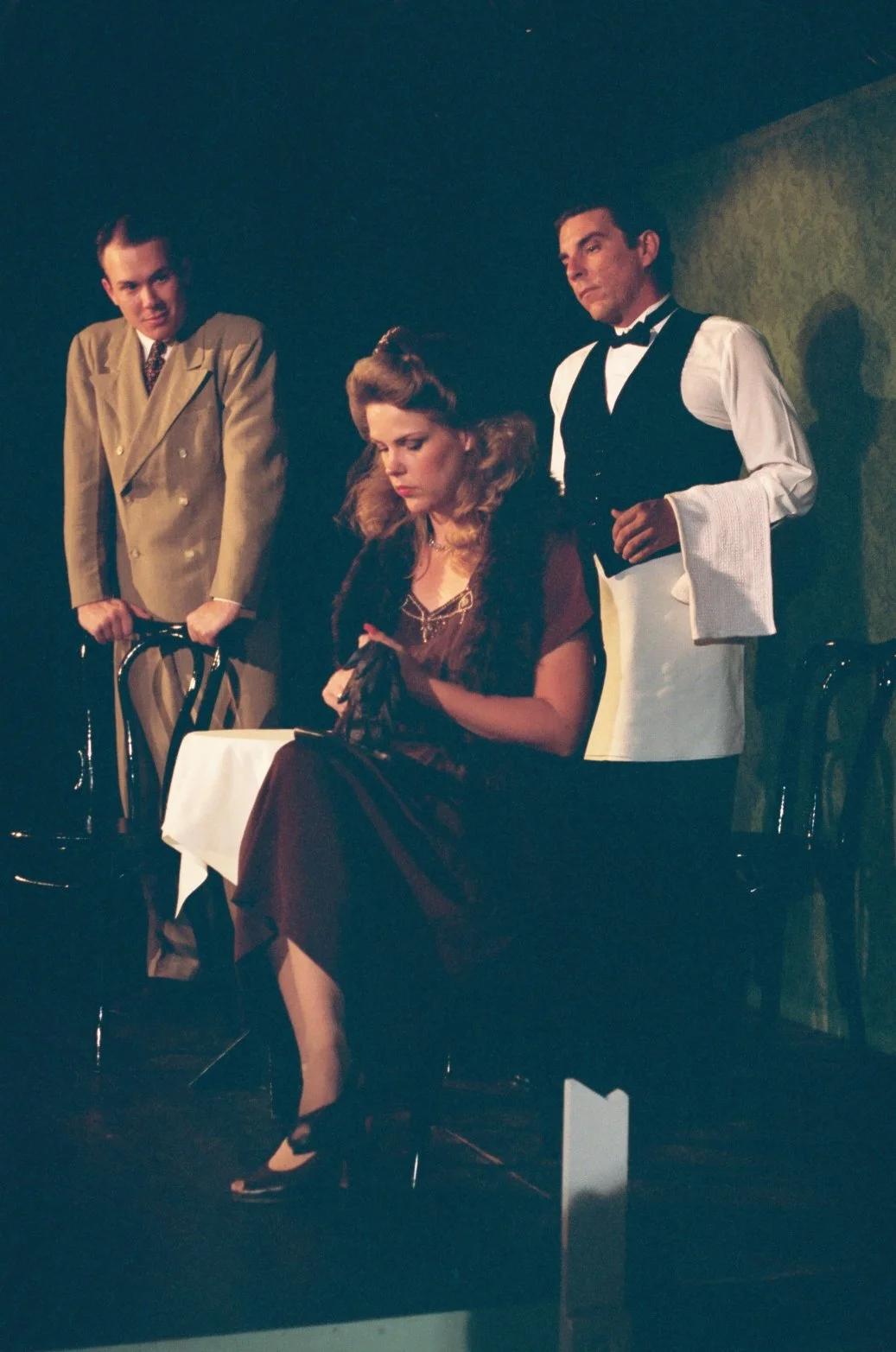 A theatrical performance with three actors: a woman seated at a table, a man in a suit leaning on a chair, and a waiter holding a towel, standing in a dimly lit setting.