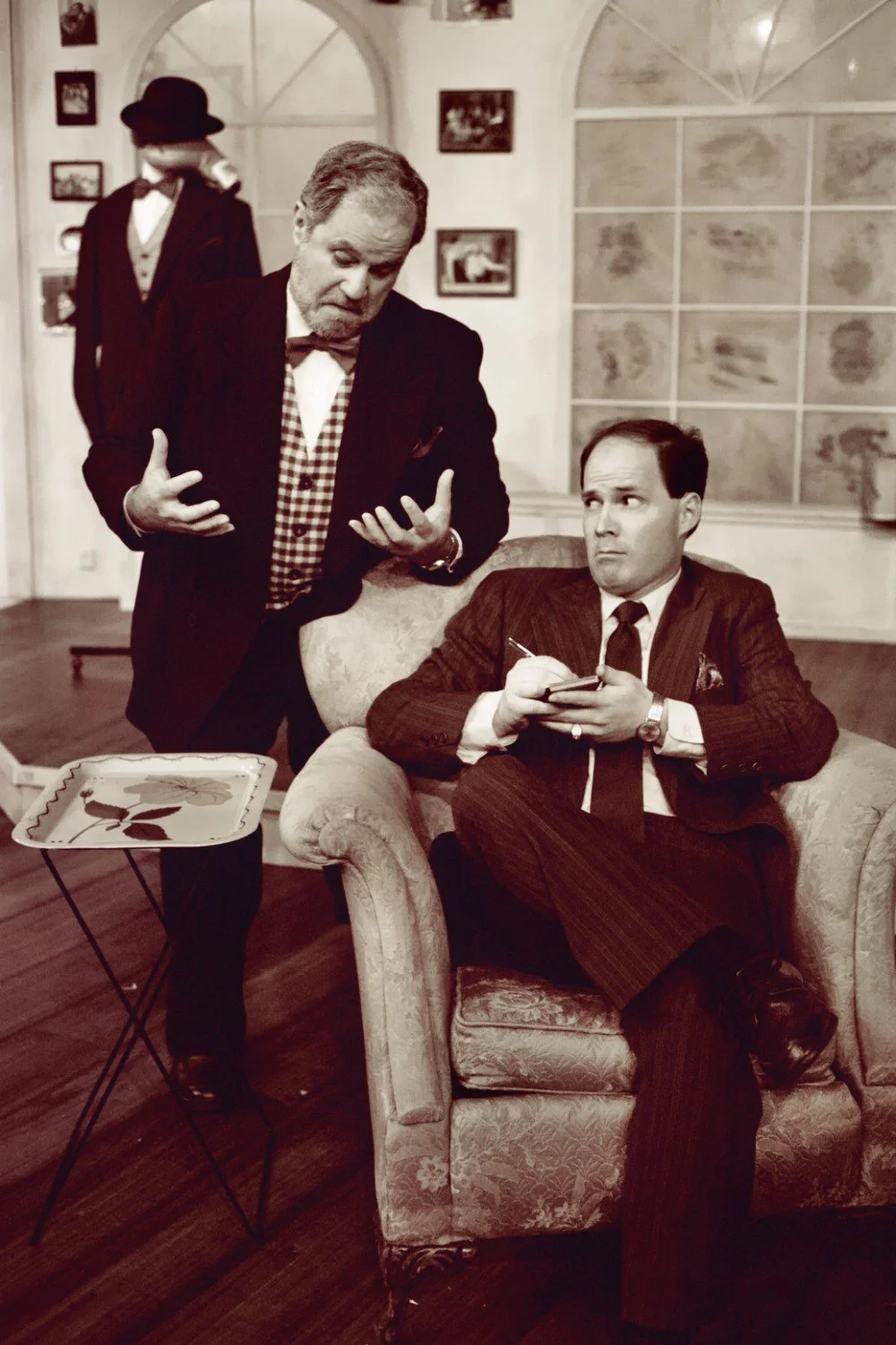 Two men in vintage suits having a conversation in a vintage-styled room, one seated in a patterned armchair with a notepad, the other gesturing with his hands. A folding tray and mannequin in the background.