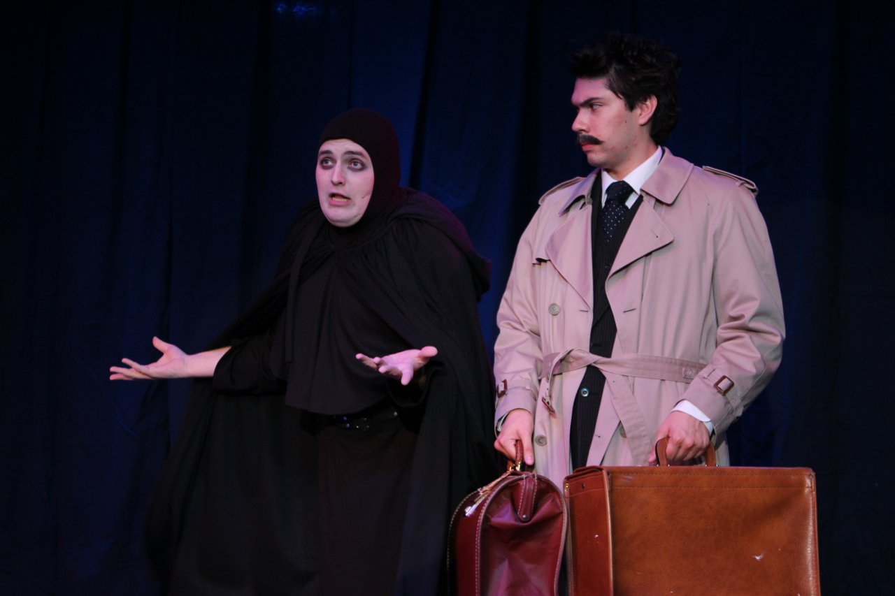 Two actors on stage, one in a black cloak gesturing with raised hands, and the other in a trench coat holding suitcases.