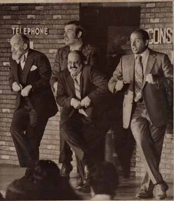 Four men performing a comedic dance routine in front of a brick wall, dressed in suits.