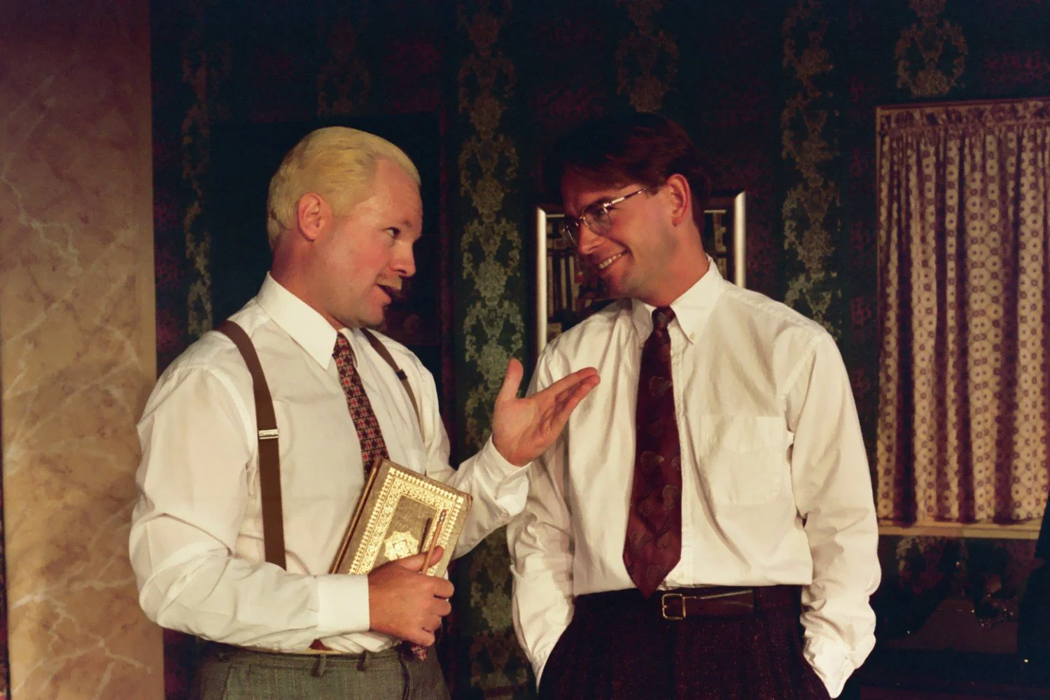 Two men in a room having a conversation, wearing white shirts and ties, one holding a book, with patterned wallpaper and curtains in the background.