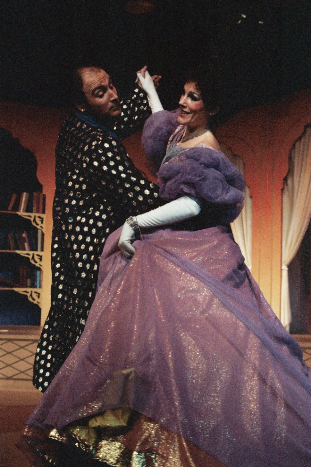 Two performers in theatrical attire dancing on stage, with the man in a patterned suit and the woman in a sparkly purple gown, in front of a set with bookshelves.
