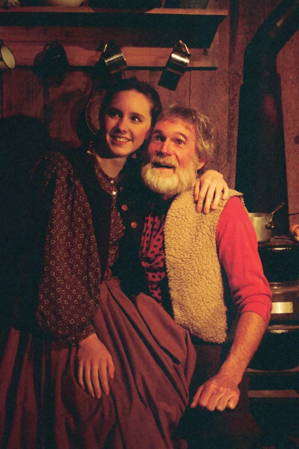 A bearded man and a young woman dressed in period costumes, sitting together in a rustic setting. The man wears a red shirt and fur vest, while the woman wears a patterned dress. They are near a wood-burning stove, with a shelf and cookware in the ba