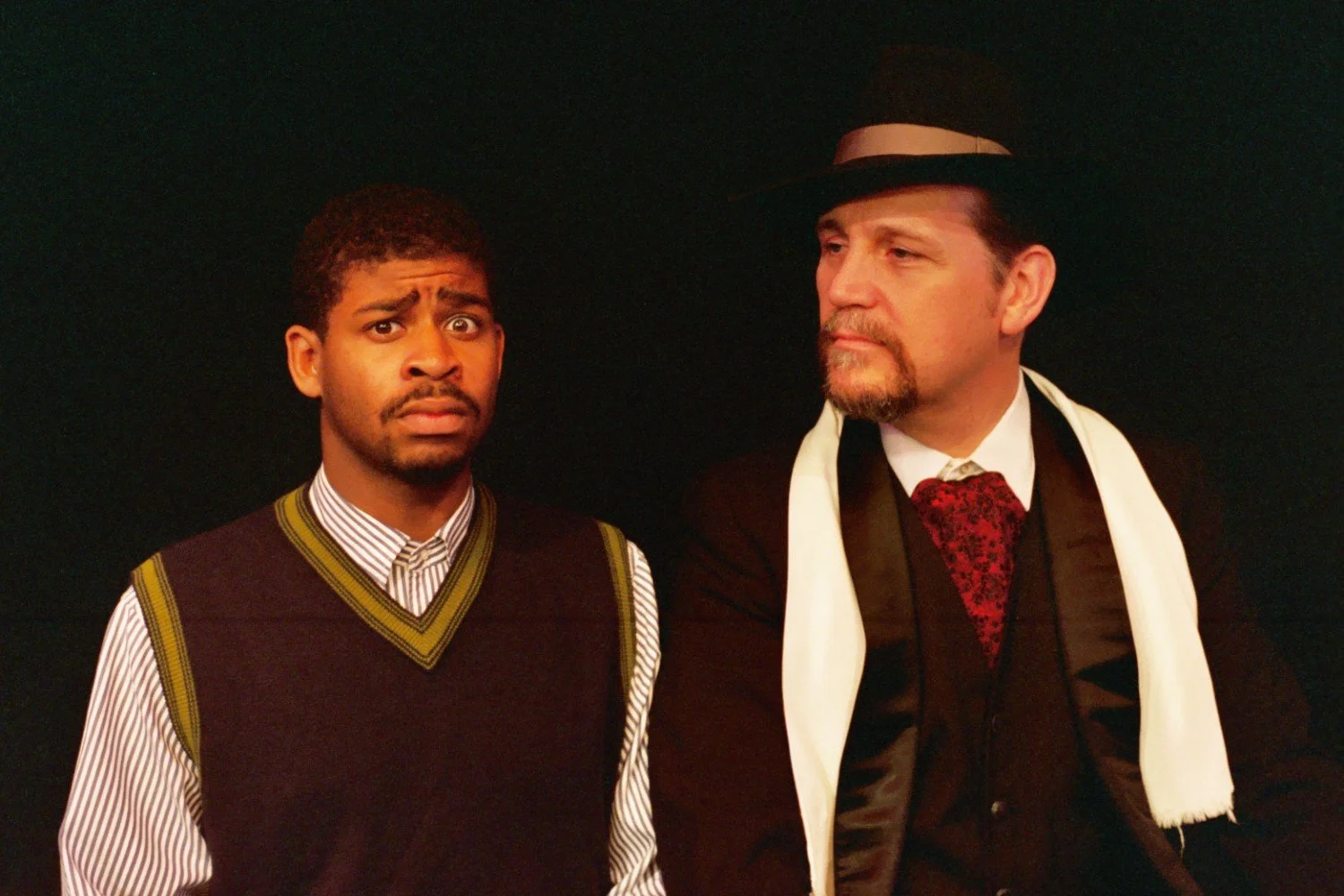 Two men in contrasting attire: one in a striped shirt and sweater vest with a concerned expression, the other in formal wear with a hat and scarf, looking calm.