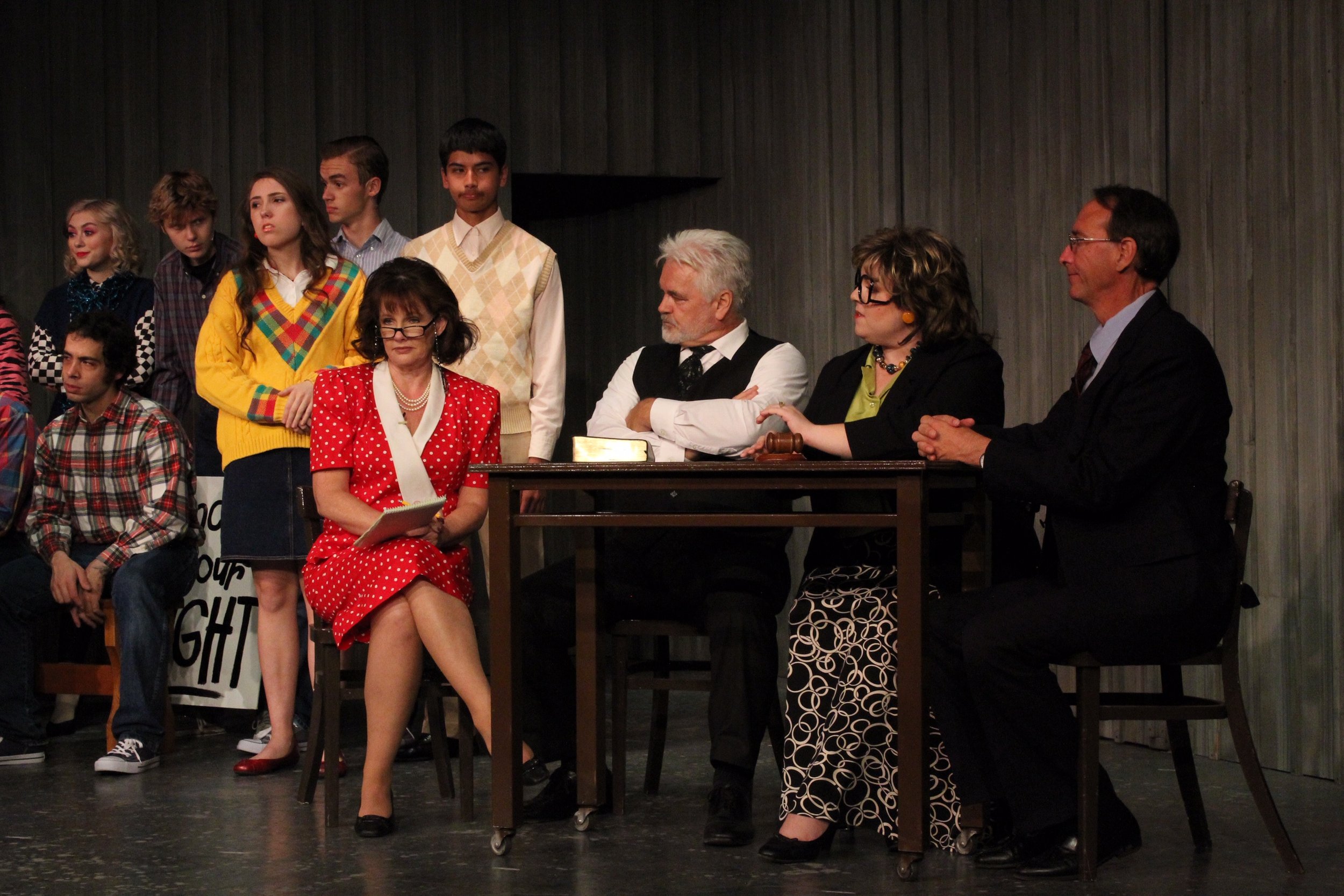 A group of people participating in a theater performance. They are wearing a mix of formal and casual vintage-style clothing and are positioned around a table on stage, suggesting a courtroom or meeting scene. Some are seated, while others are standi