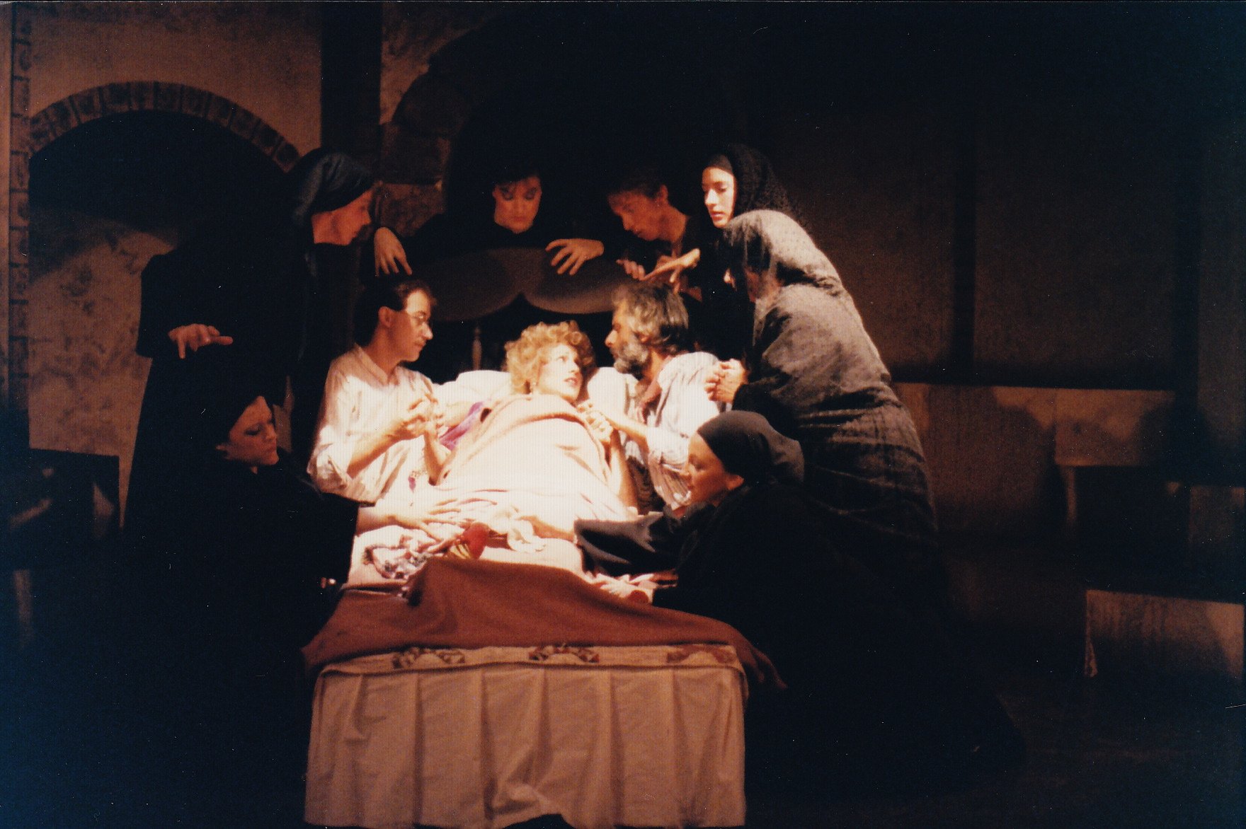 Stage scene with a group of people dressed in dark clothing gathered around a person lying in bed, appearing to be a dramatic performance.