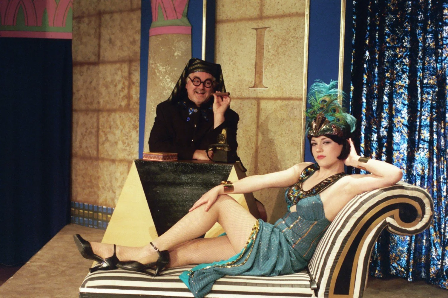A person in elaborate attire lounges on a striped chaise, while another person in a suit and glasses sits nearby holding a cigar. The setting is theatrical with a vintage style.