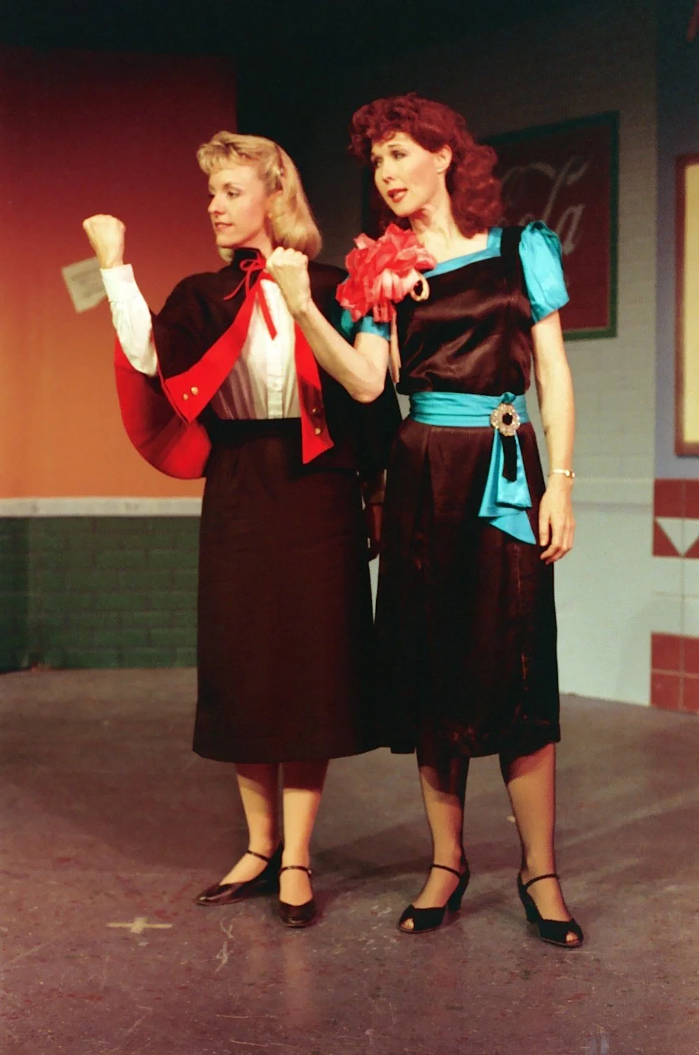 Two women in vintage-style dresses, one in red and black, the other in blue and black, on a stage with retro decor, gesturing expressively.