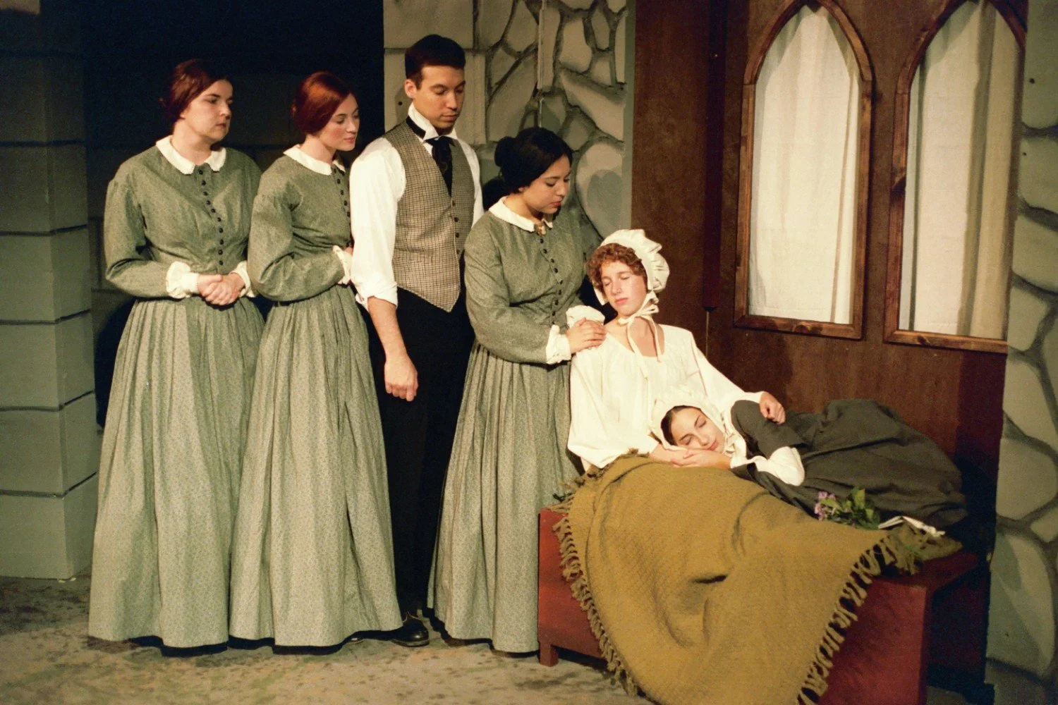 Group of people in period costumes performing a play, with women in long dresses and a man in a vest, surrounding two women on a bench, one appearing to be sleeping.