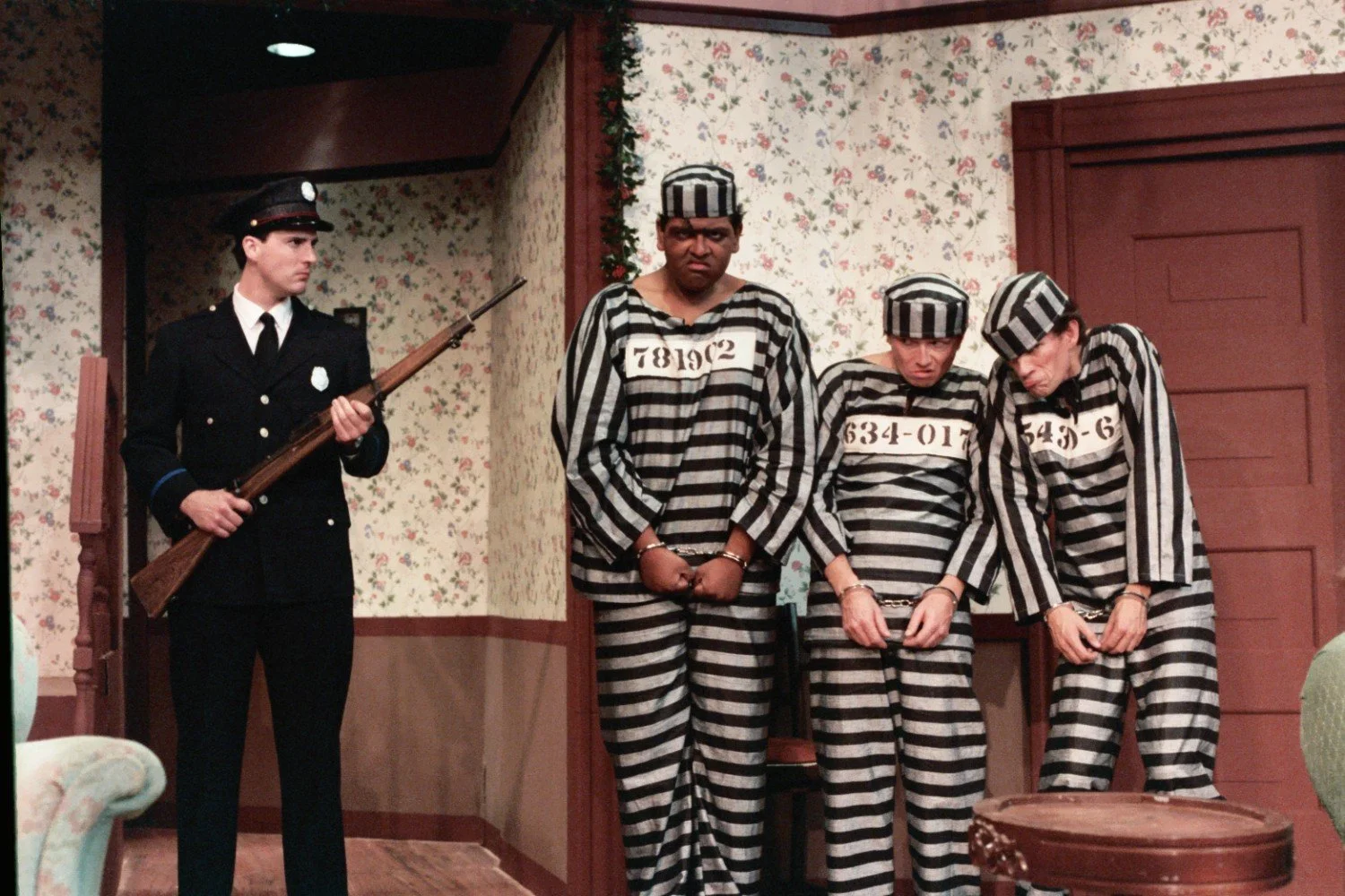 A stage scene from a play featuring a uniformed guard with a rifle standing next to three people in black and white striped prison uniforms and handcuffs in a room with floral wallpaper.