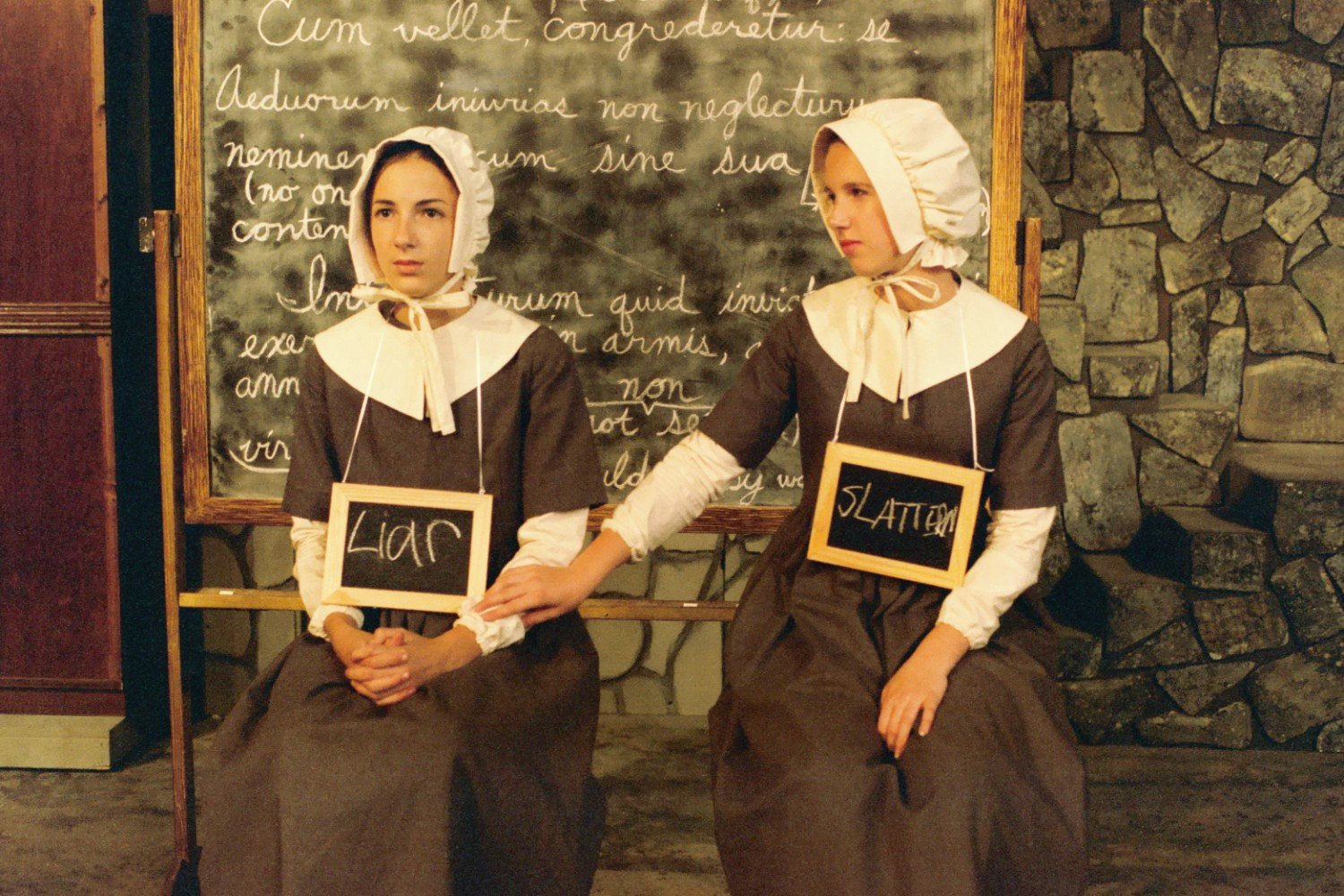 Two women in colonial-style dresses and bonnets, seated, each with a chalkboard sign around their necks reading "Liar" and "Slattern" respectively, against a stone and chalkboard backdrop.