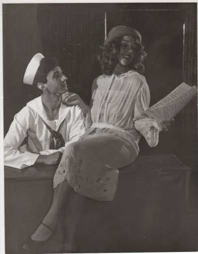 A black and white photo featuring a man in a sailor outfit and a woman in vintage clothing. The woman is holding a rolled paper and sitting on a ledge, while the man looks at her.