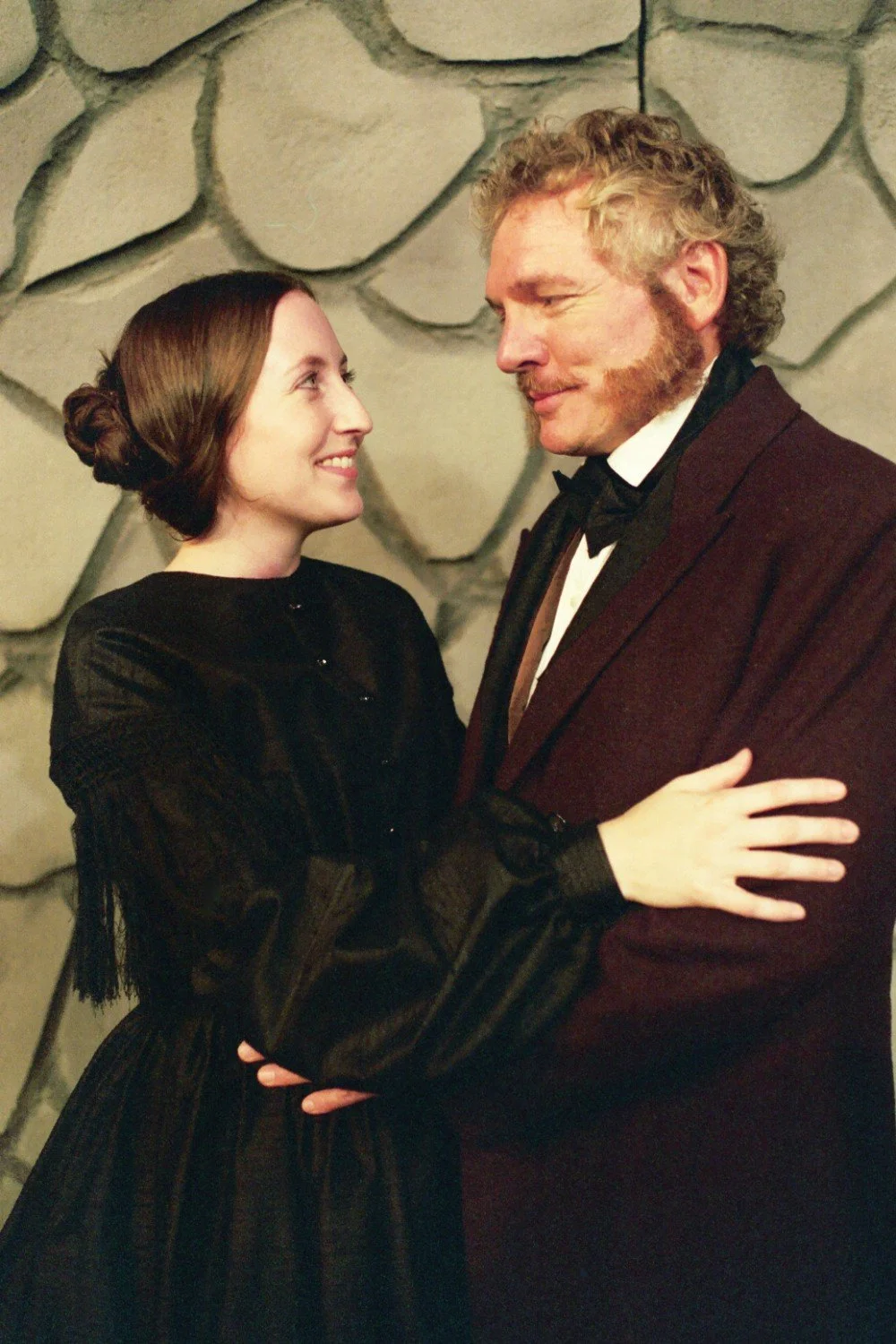 A man and woman in historical costumes embracing, with a stone wall in the background, smiling at each other.