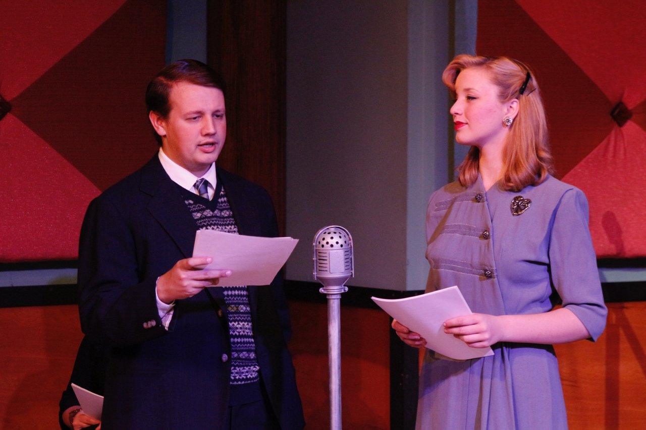 Two people dressed in vintage clothing standing in front of an old-fashioned microphone, reading from scripts during a radio performance.