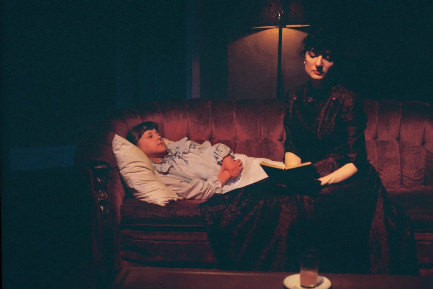 A dimly lit scene showing a woman sitting on a sofa, reading a book, while a child lies beside her with a pillow.