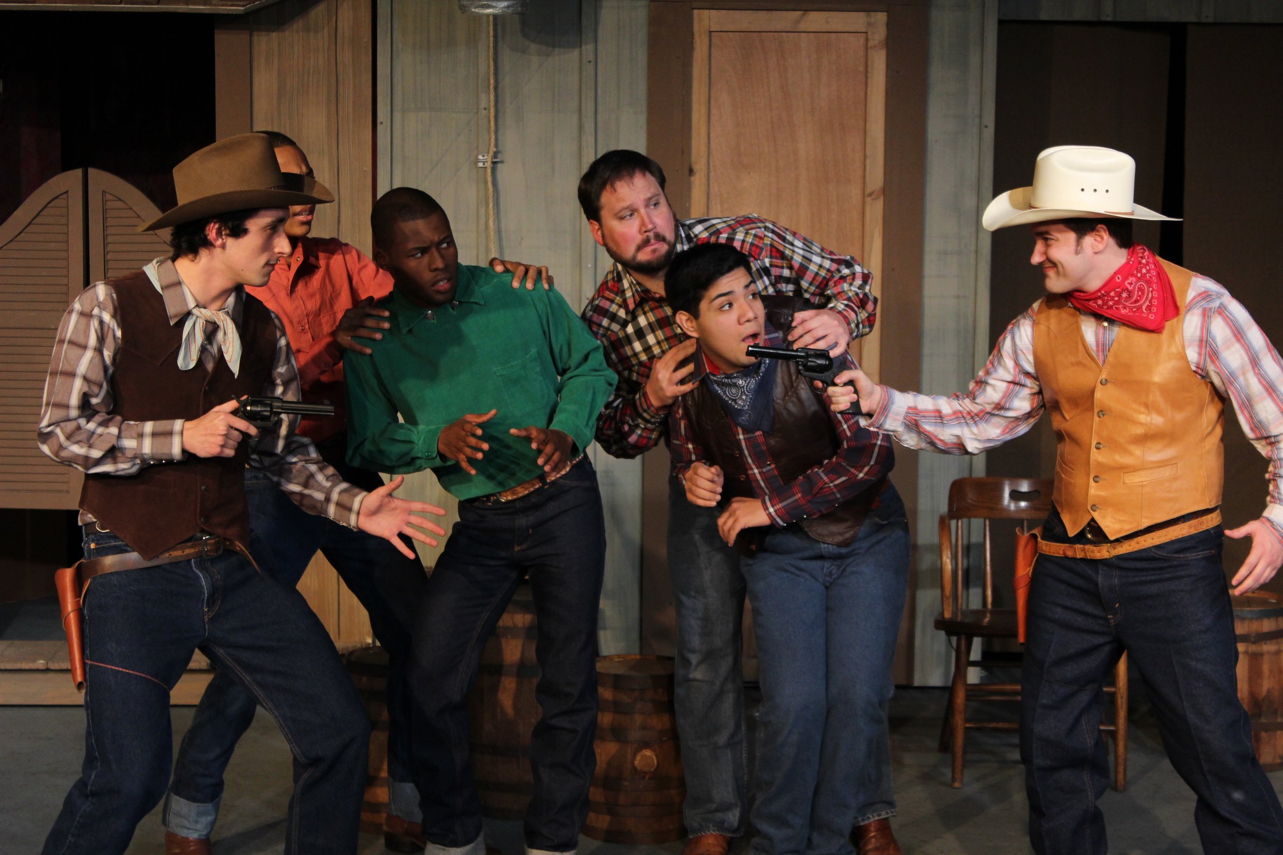Group of actors in cowboy outfits performing a humorous Western-themed scene on stage with two holding toy guns.