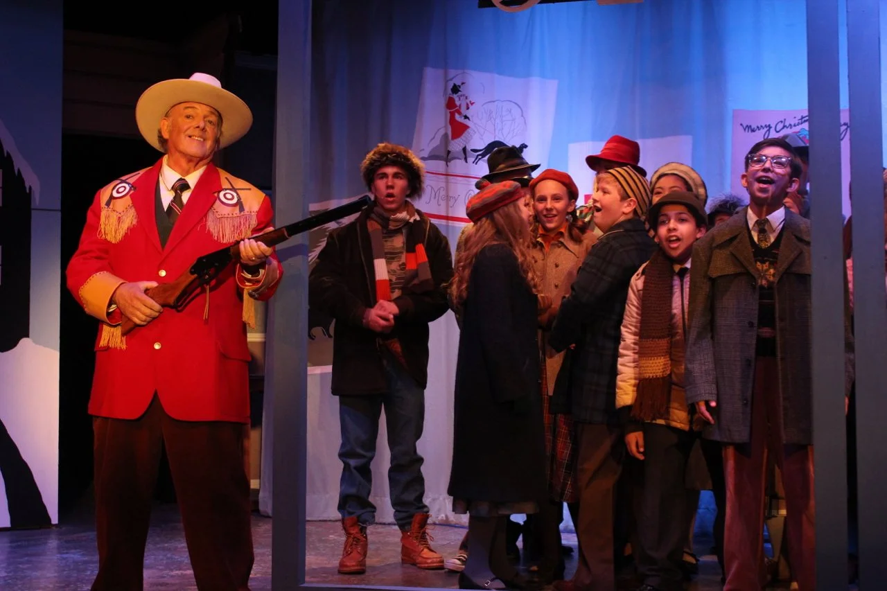 Theatre scene from "A Christmas Story" with actor in red cowboy outfit holding a rifle, surrounded by young actors in winter clothing.