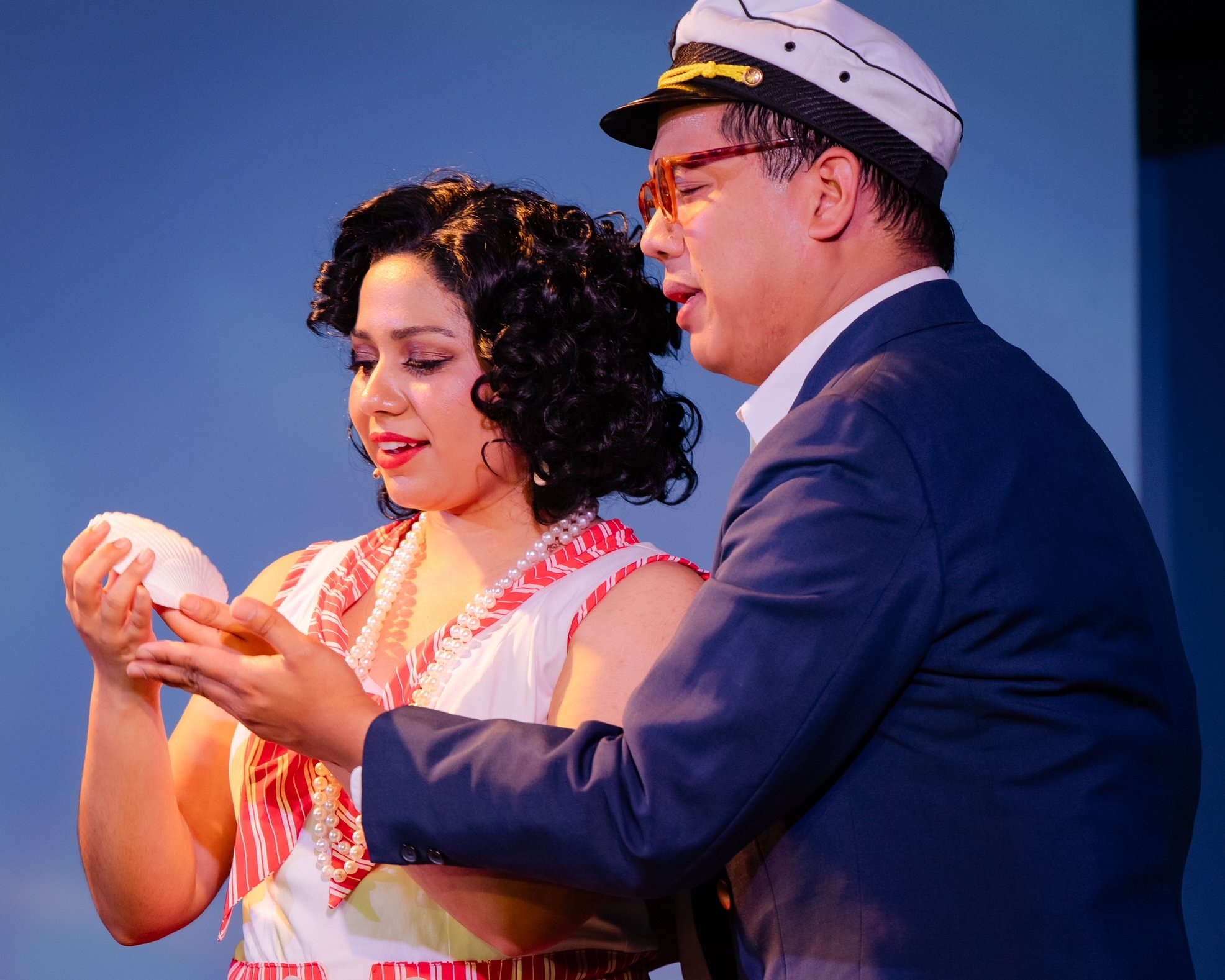 Two people on stage, one holding a seashell, the other wearing a cap.