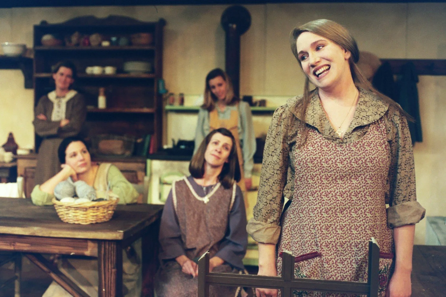 A group of women in vintage clothing inside a rustic room with a wooden table and shelves, engaged in conversation or performance.