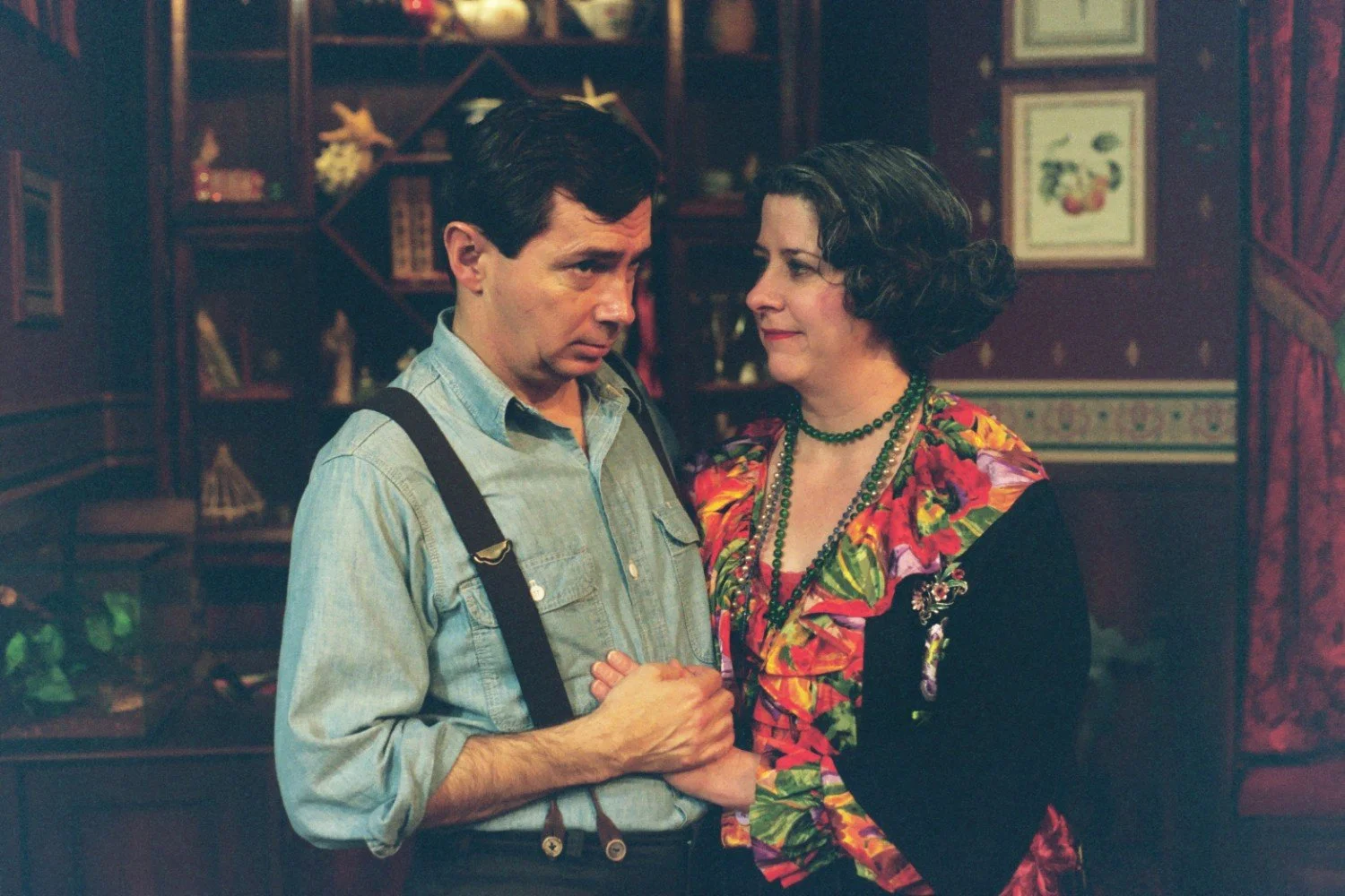 A man and woman holding hands, standing in a warmly decorated room with wooden shelves and framed art.