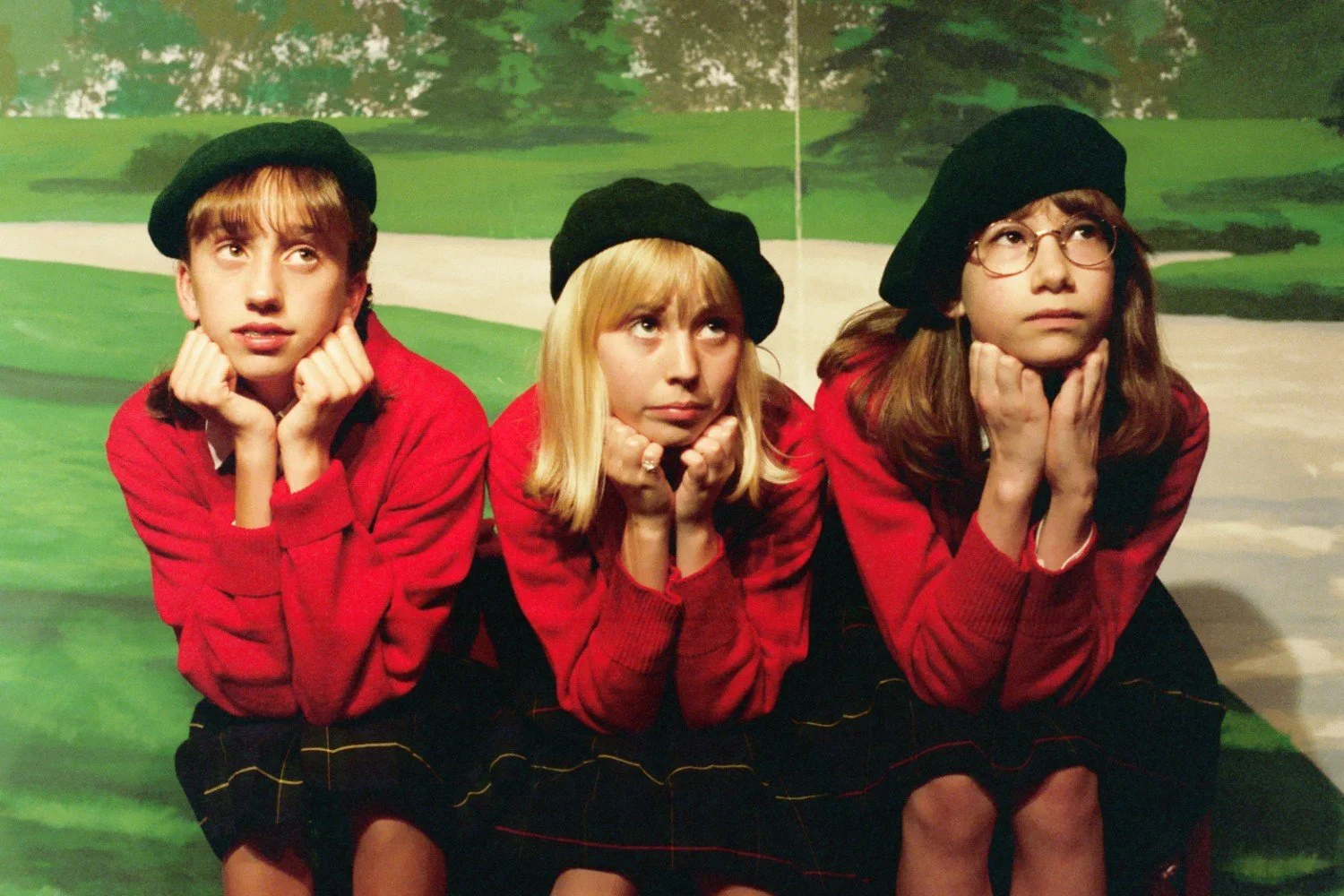 Three young people in red sweaters and black berets, sitting with their chins on their hands, looking thoughtful, in front of a painted outdoor backdrop.