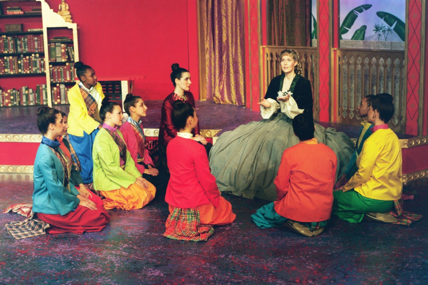 A group of children in colorful traditional outfits kneeling around a woman in a gray dress on stage, with a red and gold set background.