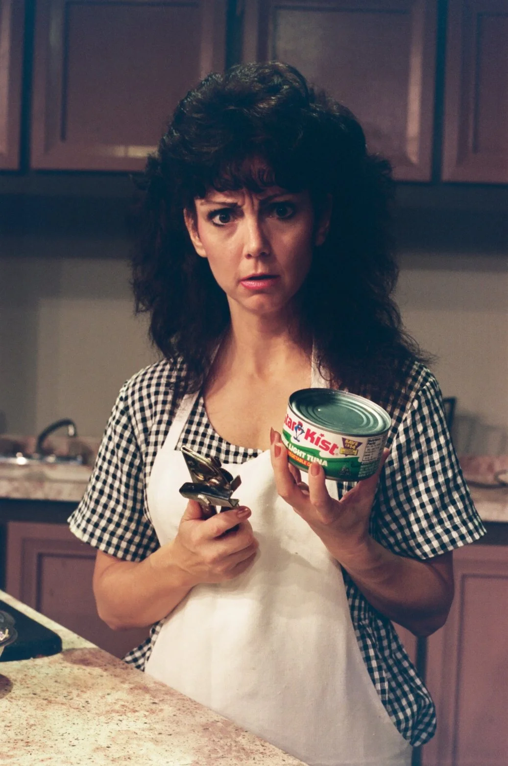 Woman holding a can of tuna and a can opener in a kitchen, looking confused.