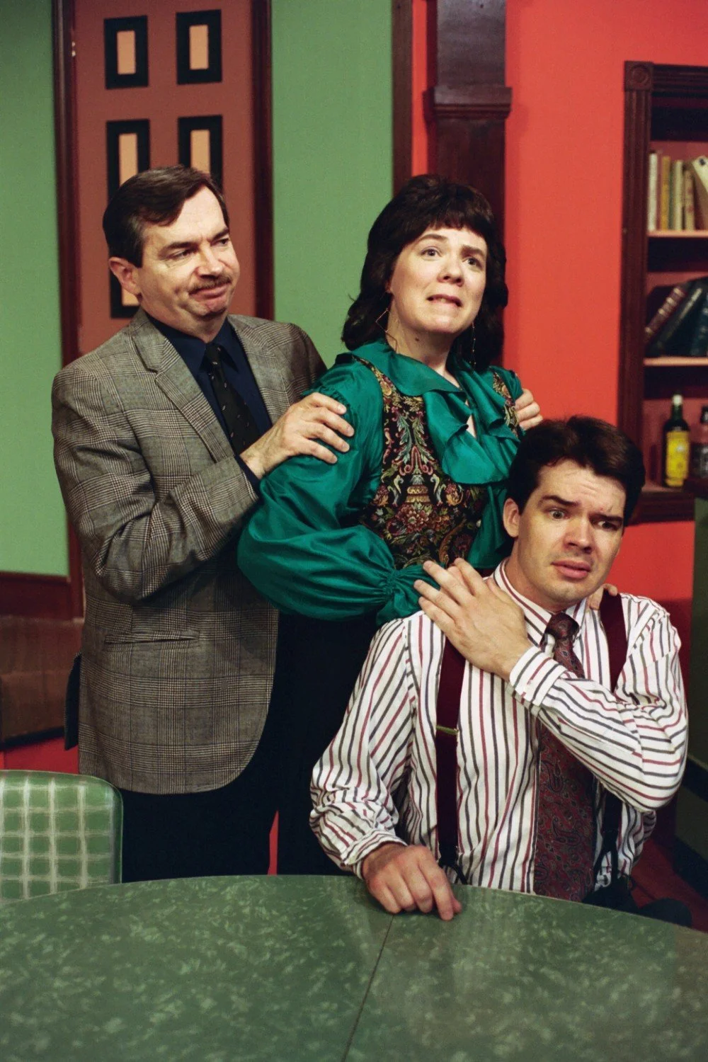 Three people dressed in vintage-style clothing standing in a room. The man on the left wears a plaid blazer, the woman in the middle wears a green and patterned dress, and the man on the right wears a striped shirt with suspenders. They appear to be 