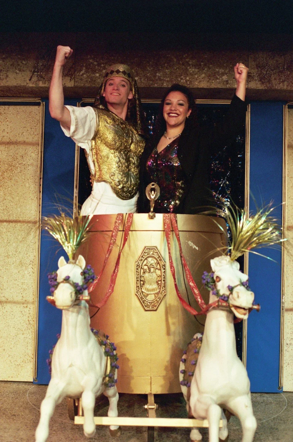 Two people posing in a decorative chariot, wearing costumes; one is dressed in a gold warrior-like outfit and the other in a sparkling dress. Two white horse sculptures with flowers are at the front of the chariot.