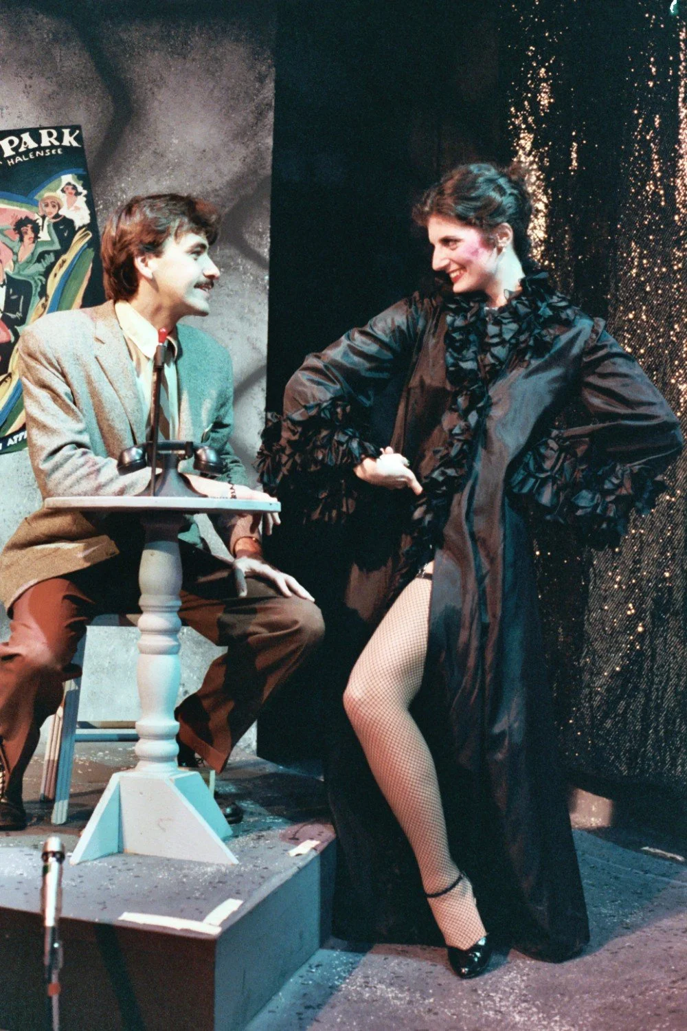 Two performers in a theatrical setting, one sitting and one standing in a playful pose, with a microphone and a vintage poster in the background.
