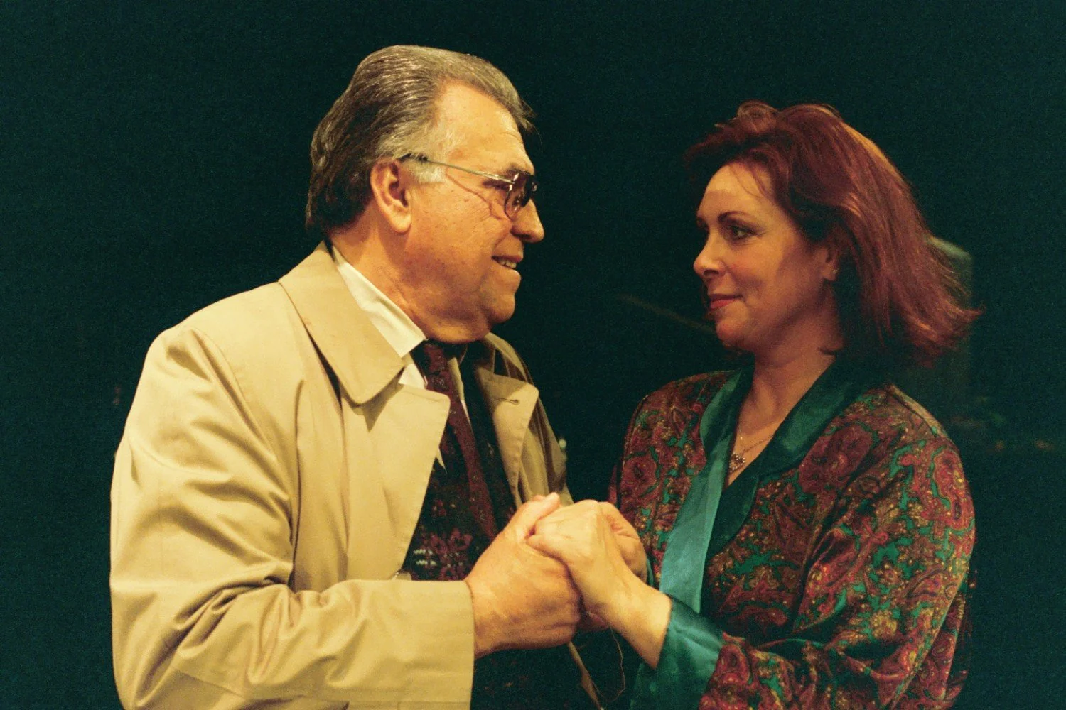 An older man in a beige trench coat holding hands with a woman in a patterned robe, looking at each other affectionately against a dark background.