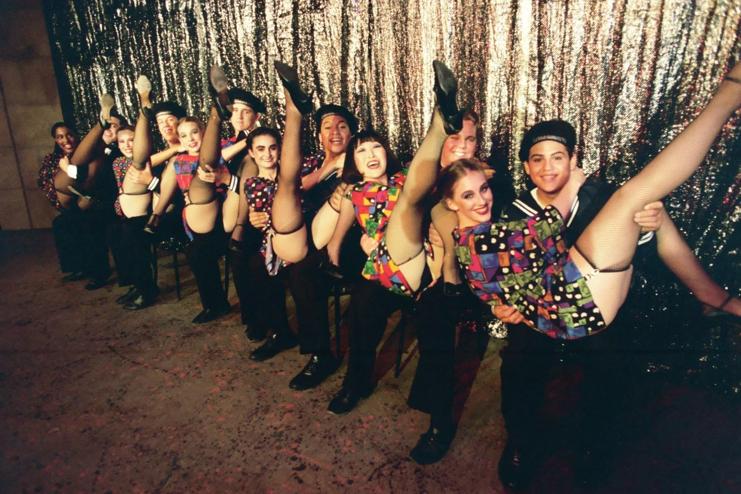 Group of performers in colorful costumes and sailor hats doing a kick line in front of a glittery curtain.