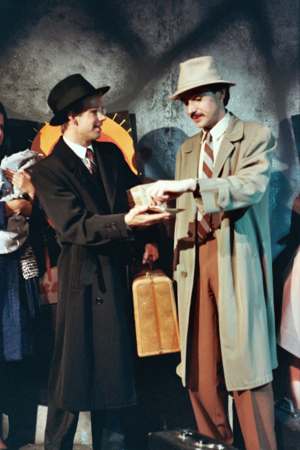 Two men in vintage attire exchanging money, one holding a suitcase, in a theatrical setting.