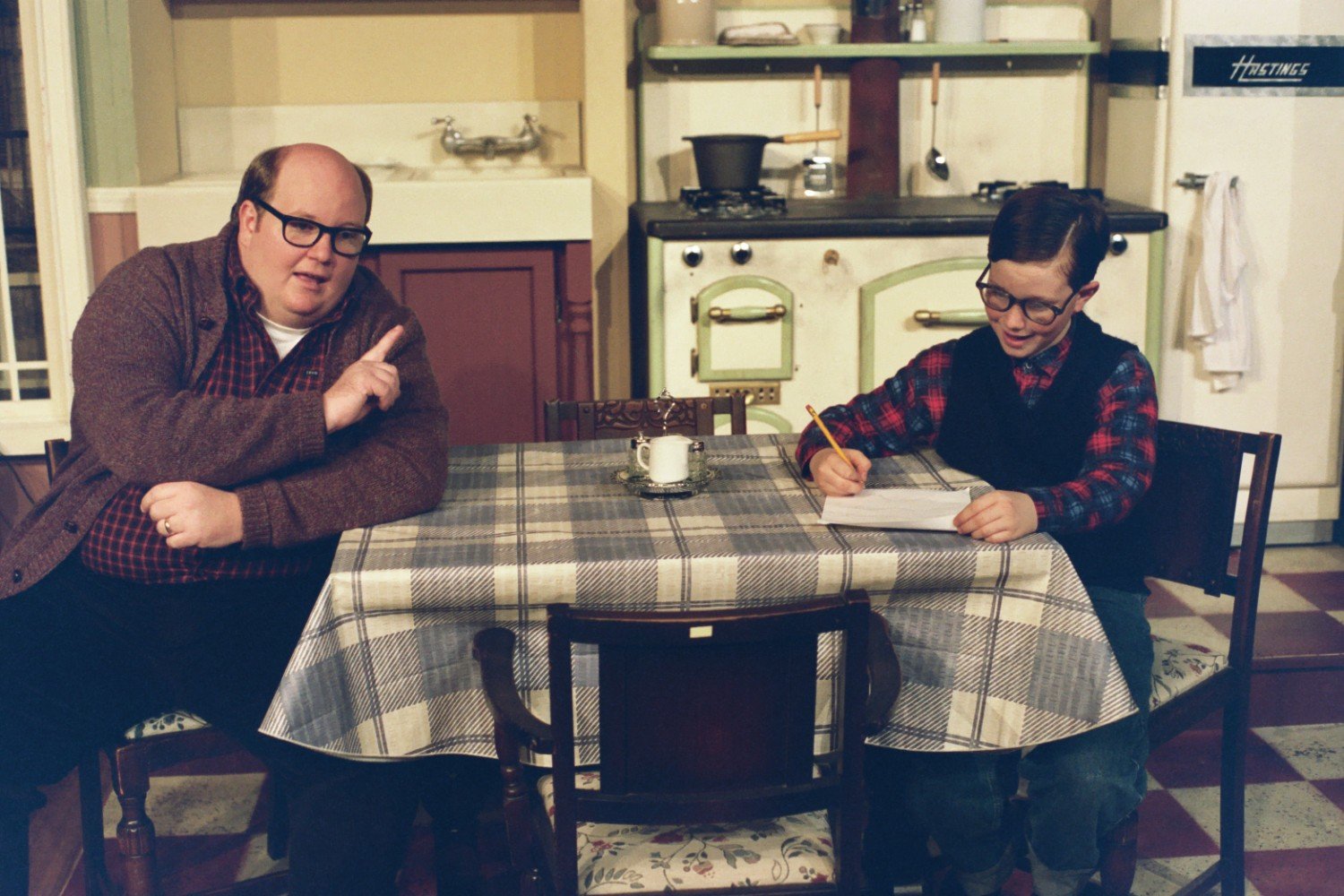 A man and a child sitting at a kitchen table with a plaid tablecloth. The man is gesturing with his finger while the child writes with a pencil. The background includes a sink, stove, and refrigerator.