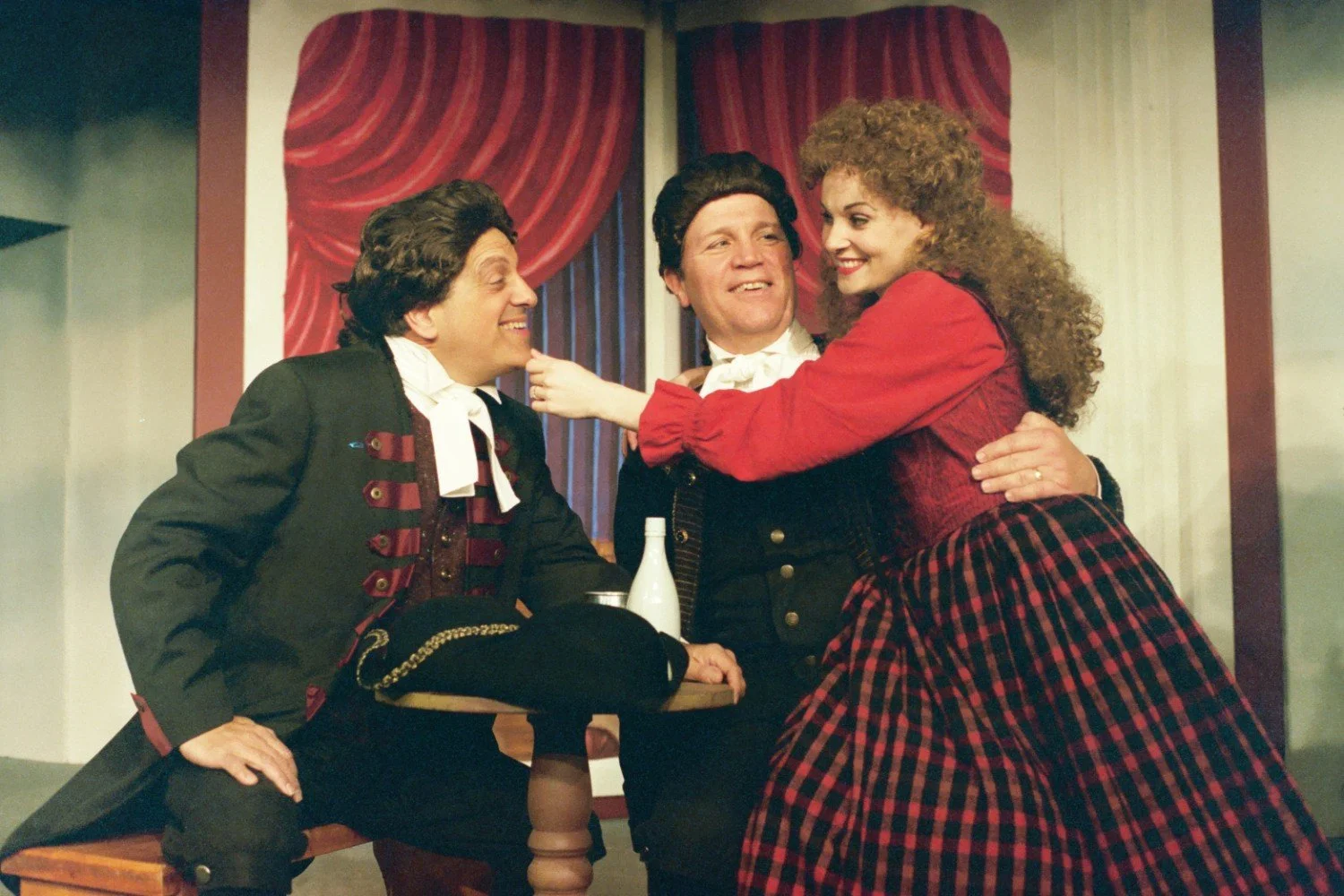 Three actors in period costumes perform on stage, featuring two men and a woman in 18th-century attire with wigs, against a backdrop of red drapes. The woman playfully touches one man's chin while they all smile.
