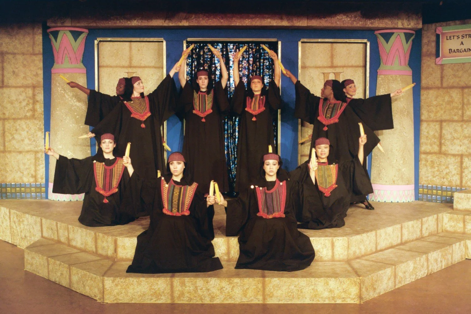 Theatrical performance with actors in black robes holding scrolls on a stage with columns.