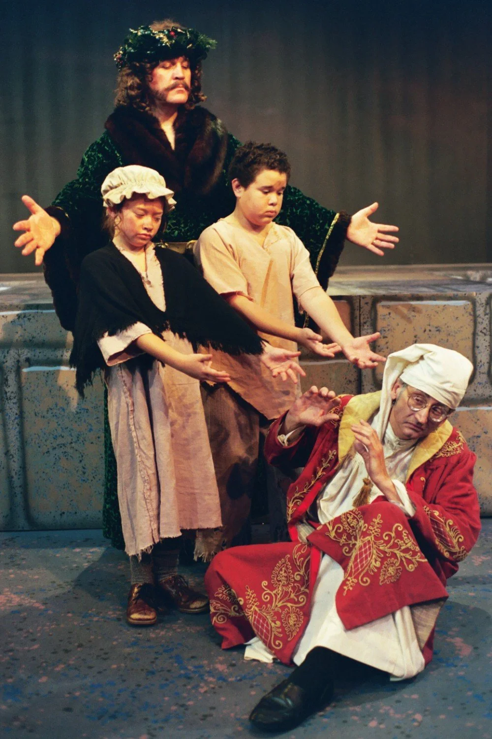 Theatrical scene with four actors in costume depicting a dramatic moment. One actor sits on the ground in an ornate red robe and white nightcap, looking frightened. Behind him, three actors stand: a man with a green robe and wreath, and two children 