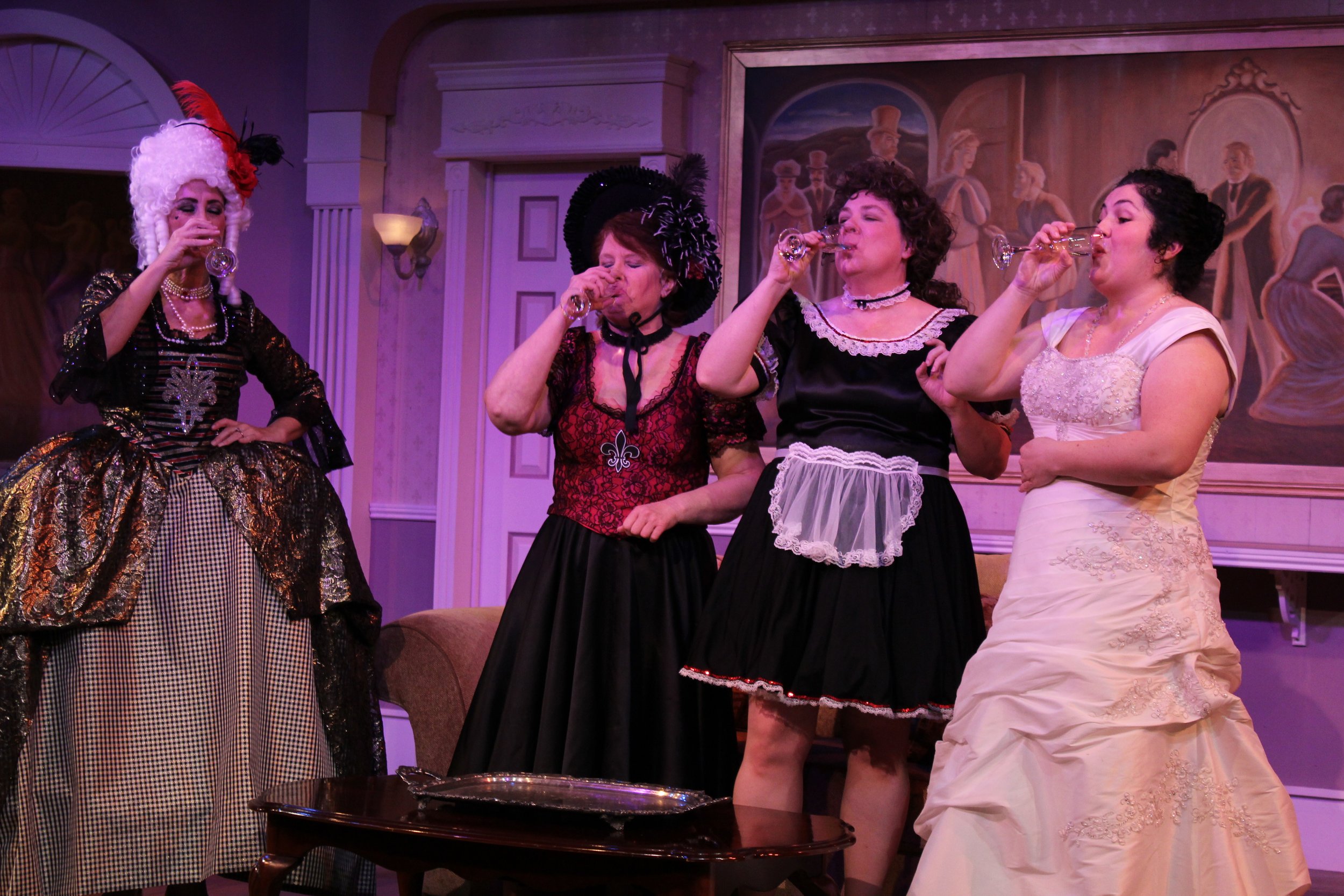 Four women in period costumes drinking from wine glasses on stage.