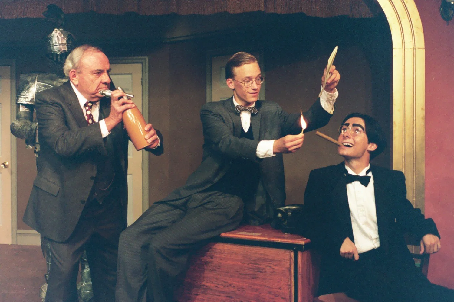 Three men in a humorous theater scene, with one holding a beverage and another lighting a match to a cigar held by the third. They are dressed in formal suits with a vintage setting.