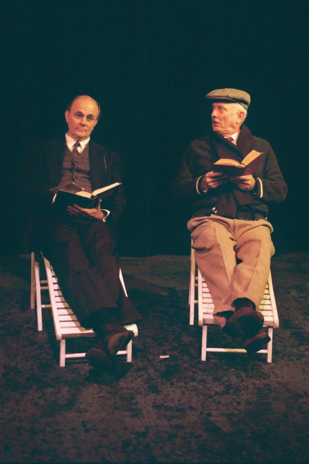 Two men sitting on chairs reading books, with one wearing a cap and scarf, in a dimly lit setting.