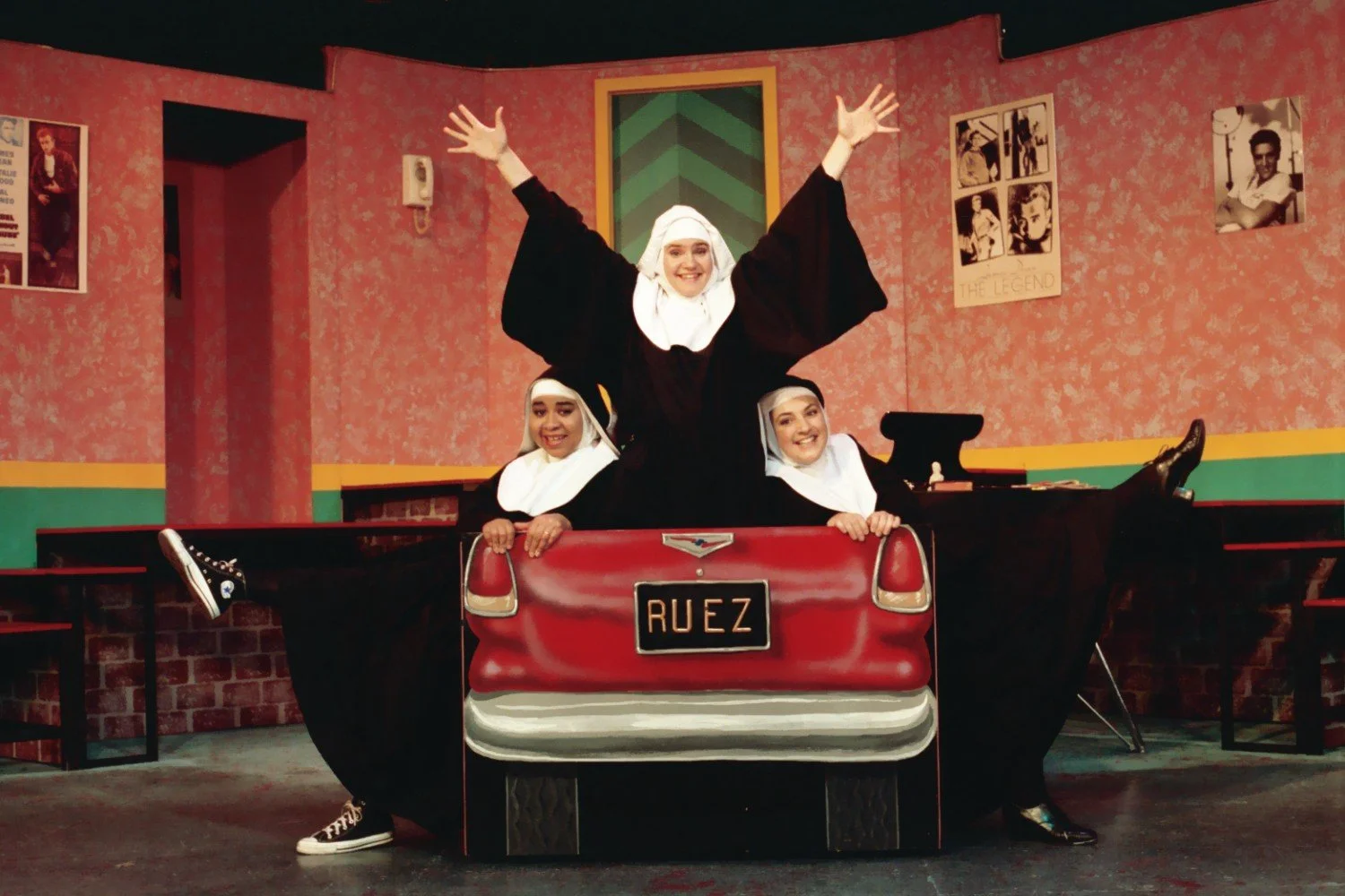 Three people dressed as nuns posing playfully in a theater scene, with a backdrop resembling a large red car and retro-style decor.