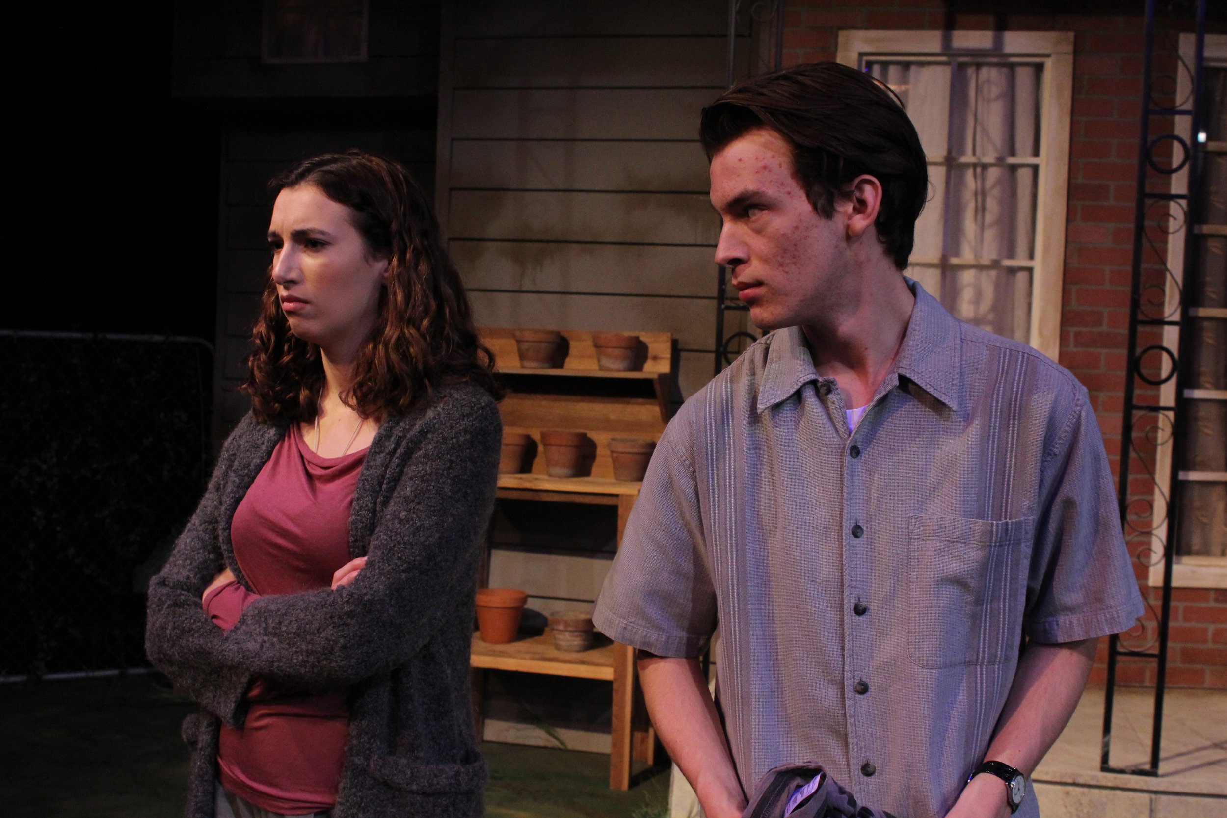 Two people standing outside near a house, with one crossing her arms and the other looking to the side, both appearing serious, in a theatrical setting.