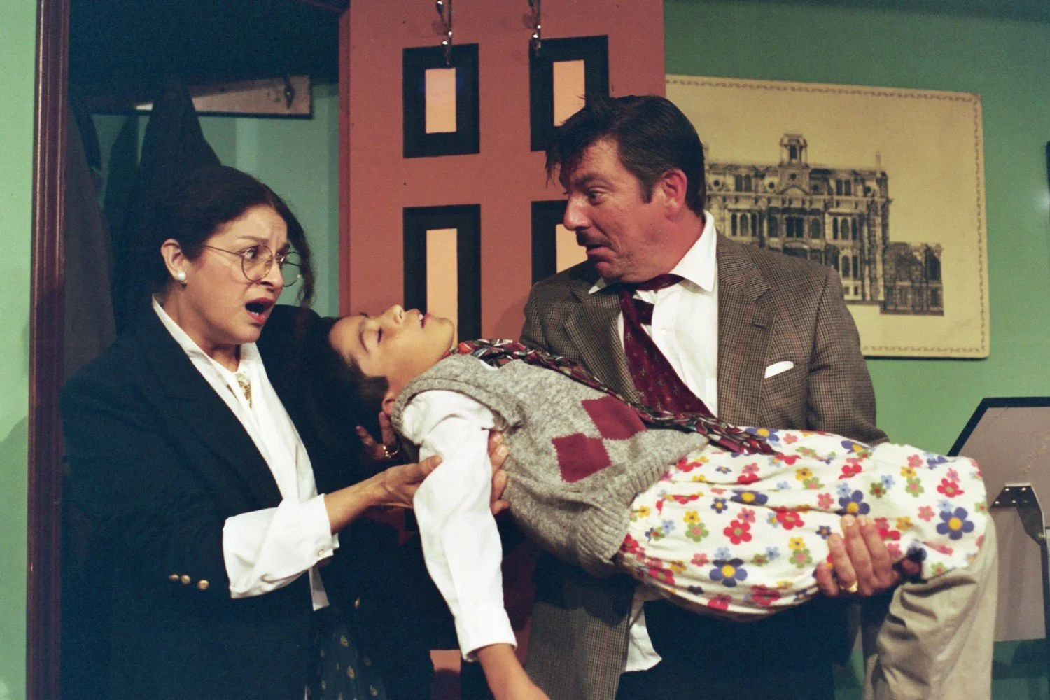 Man and woman in a theatrical setting, with the man holding an unconscious child dressed in colorful clothes. The woman is reacting with concern, and the scene is set against a backdrop of a room with a door and wall art.
