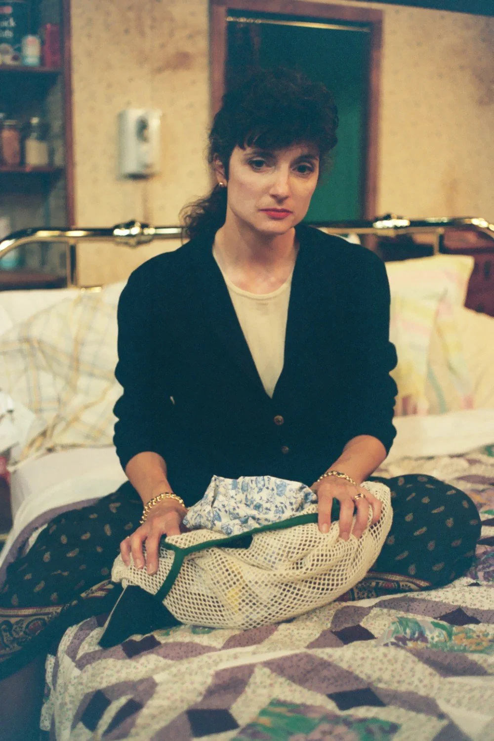 A woman sitting on a bed, holding a knitted or crocheted blanket with a floral pattern. The room has a vintage style, with patterned wallpaper and a visible shelf in the background.