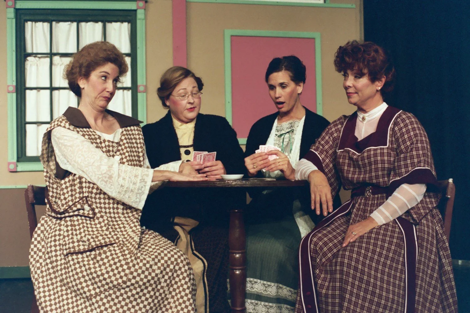 Four women in vintage costumes playing cards at a table in a theater setting.