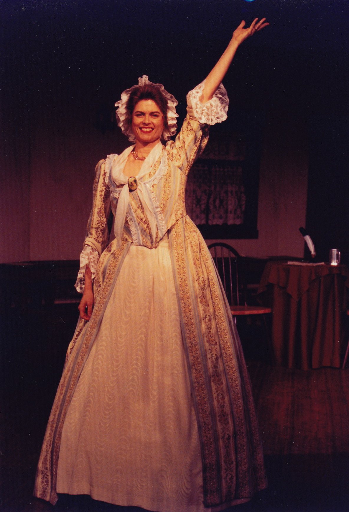 Woman in 18th-century dress on stage with arm raised.