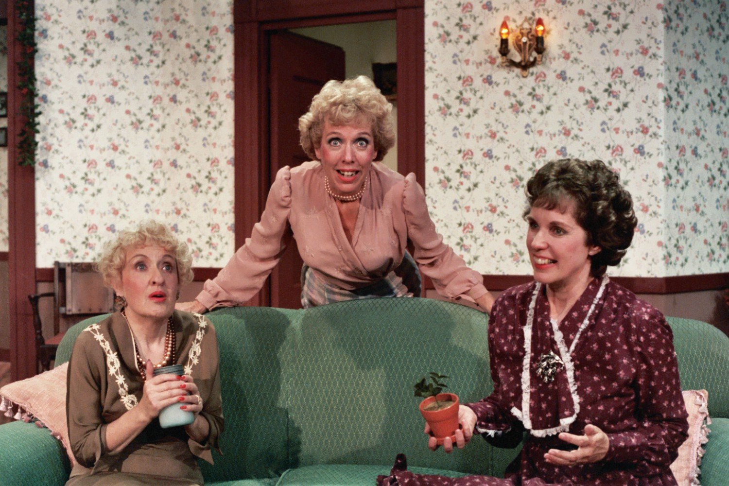 Three women in a vintage-style living room; one has curly blonde hair and an apron, another is holding a cup with both hands, and the third is holding a small potted plant.